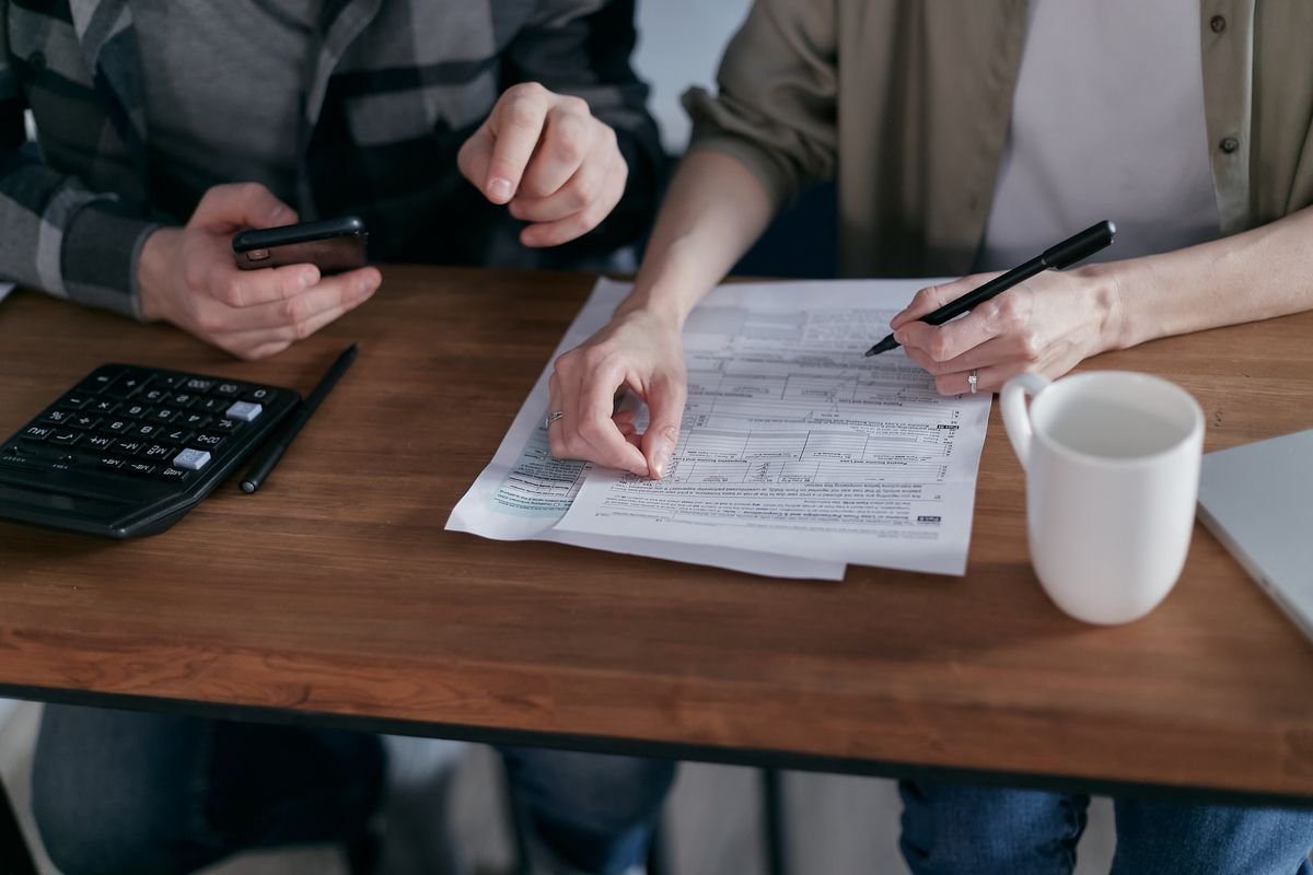 Two people at a wooden table reviewing a printed form with a pen and a phone, beside a calculator and a white mug