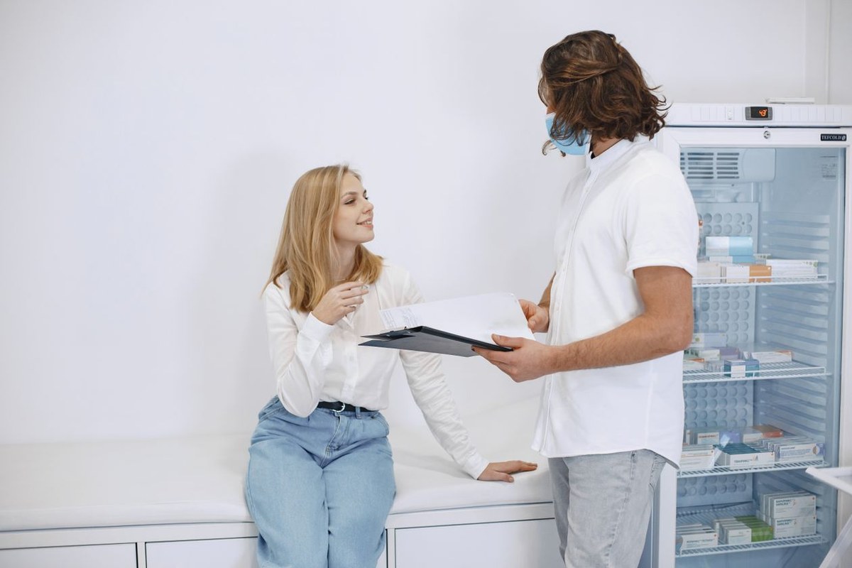 Patient on an exam bench talking with a masked clinician holding a clipboard beside a medical fridge